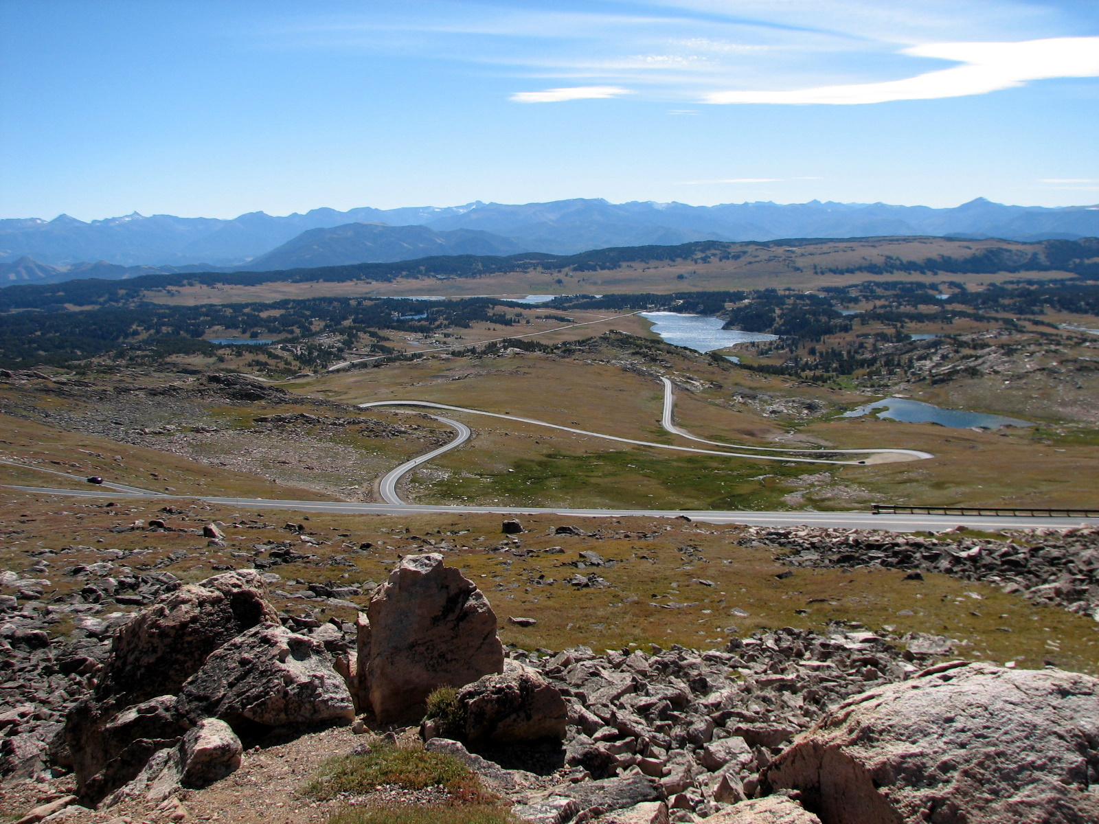 Switchback turns of the Beartooth Highway climbing the Beartooth Plateau, with snowfields visible above tree line and the Absaroka mountains stretching to the horizon.
