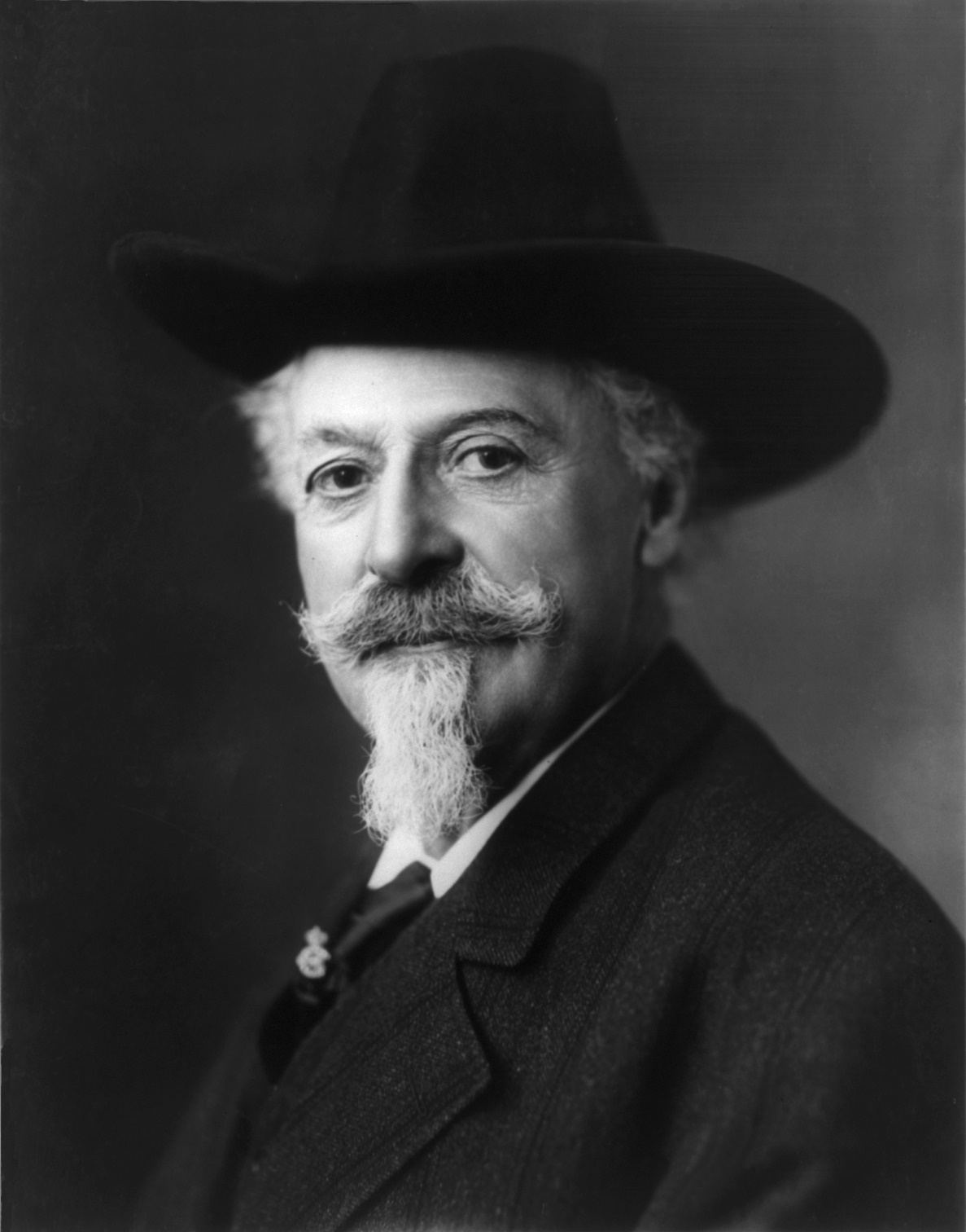 Period portrait of Buffalo Bill Cody in long hair, goatee, fringed buckskin jacket, and broad-brimmed hat, holding a Winchester rifle.