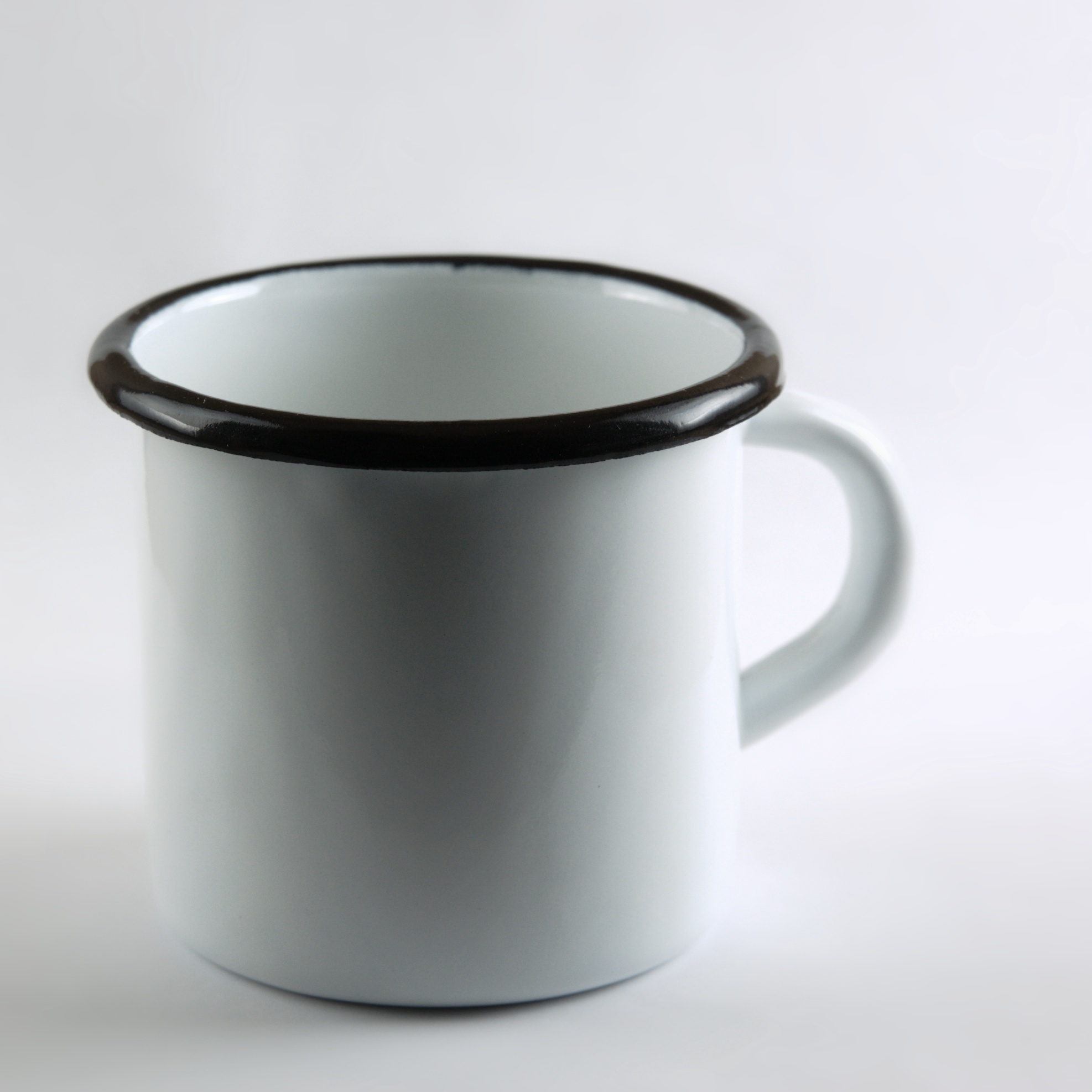 A white enamelware mug with a dark rolled steel rim, photographed against a plain light grey background, showing the smooth glassy porcelain-enamel surface and the characteristic wrapped steel edge at the lip.