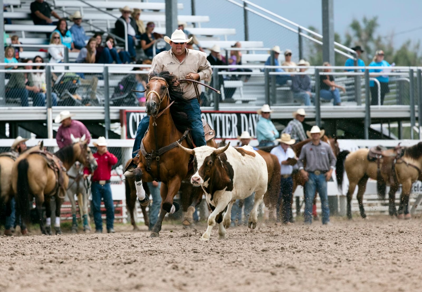 A tie-down roper chasing a calf across the Frontier Park arena, with the open aluminum bleacher sections and standing contestants visible behind the arena fence.