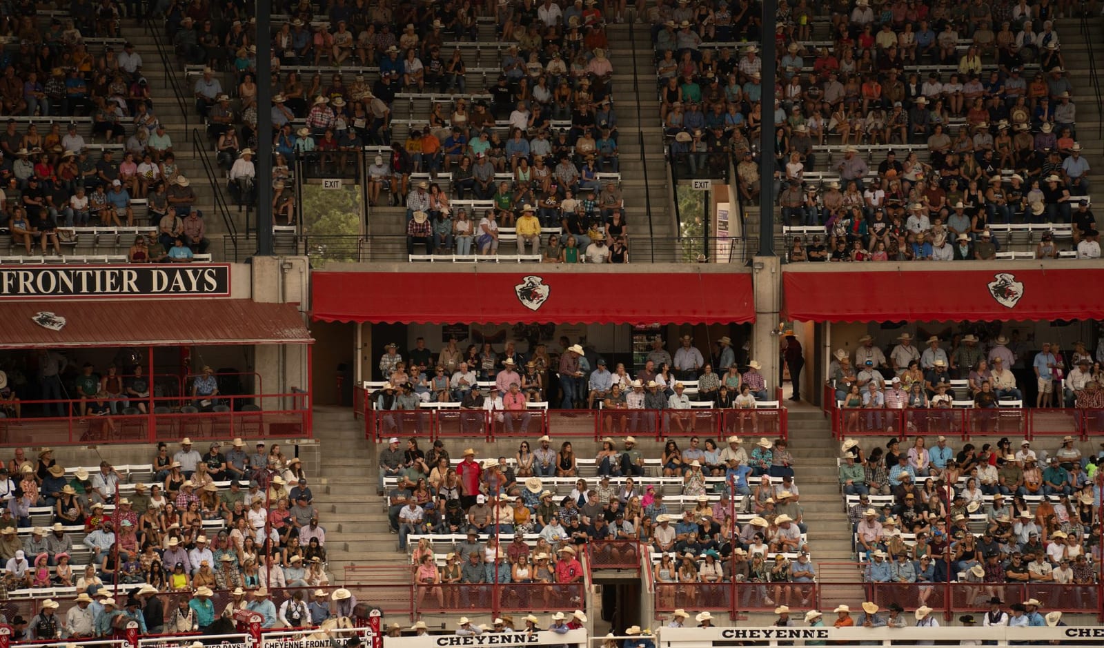 Packed grandstands at Cheyenne Frontier Days, with the covered box seats in the middle tier and open bleacher sections above and below, most of the crowd in straw hats.