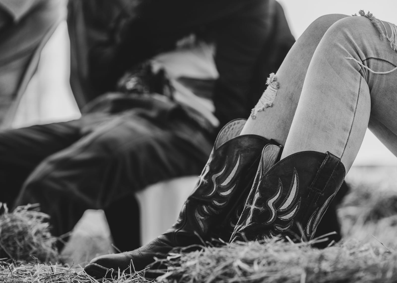 Black and white close-up of a pair of worn cowboy boots and jean legs on straw ground.