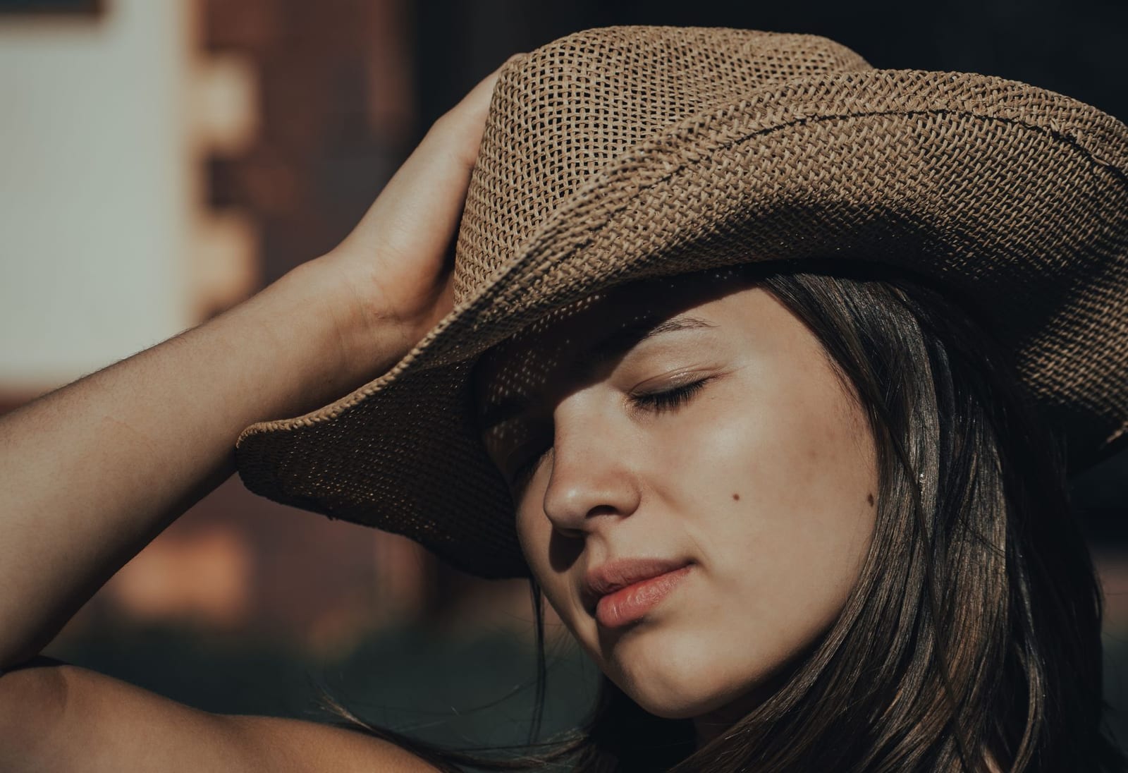 Close portrait of a woman in a wide-brimmed straw cowboy hat with her eyes closed in full sunlight, the brim casting a hard shadow across her face.