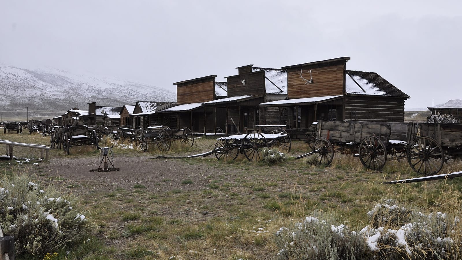 The single street of Old Trail Town in Cody, Wyoming, a row of weathered log and board frontier buildings with period wagons parked in the sagebrush in front of them, light snow on the ground.