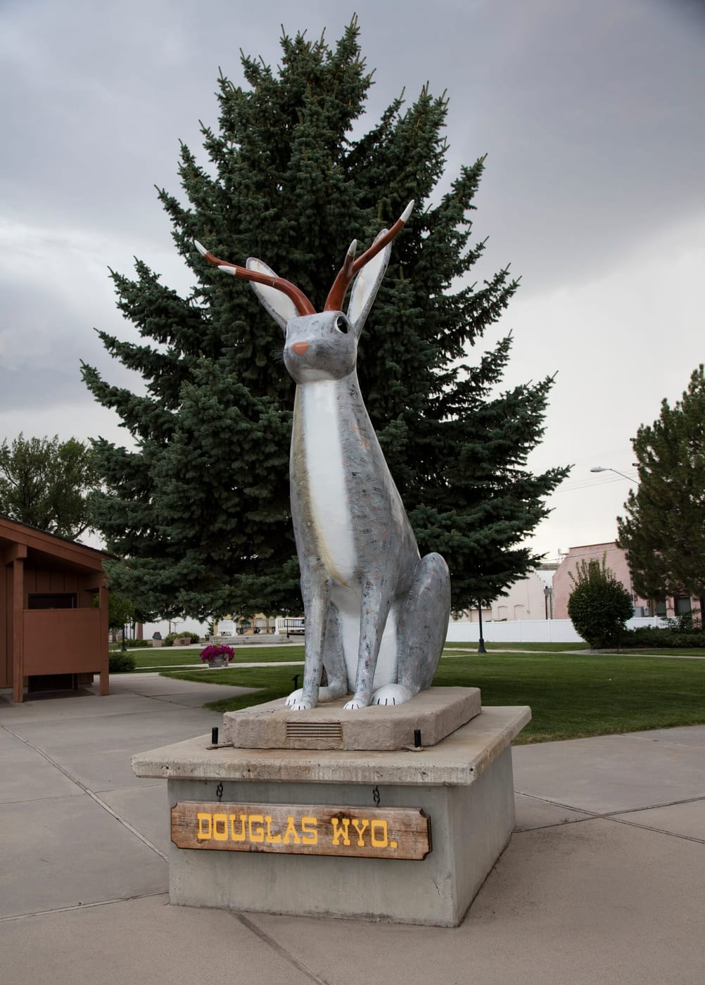 The tall fiberglass jackalope statue in Douglas, Wyoming, a seated jackrabbit with antelope antlers, standing on a plinth in a small park with pines behind it.