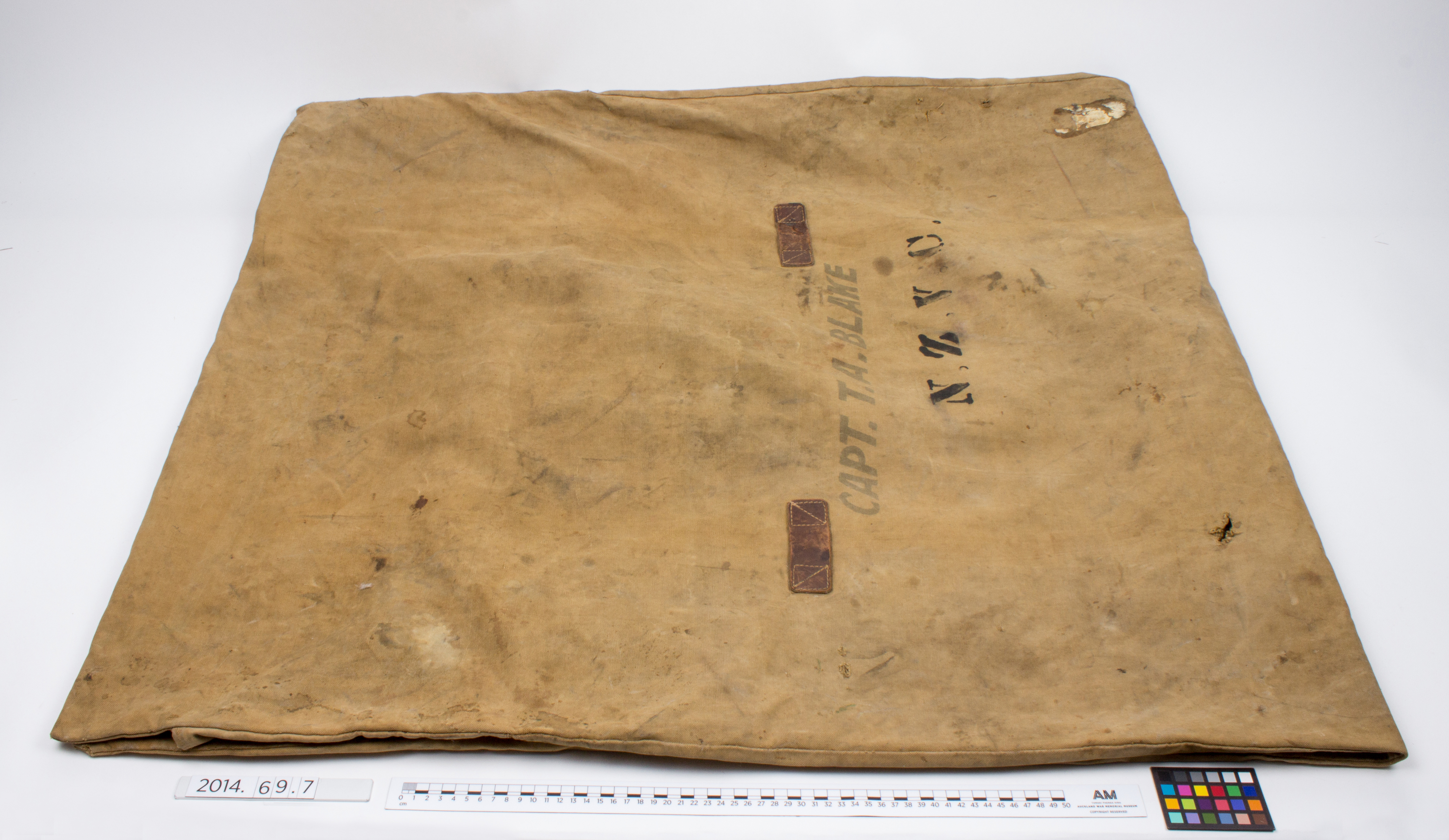 A tan canvas ground sheet and bedroll laid flat, showing two brown leather strap patches, a crossed-stitching reinforcement point, and stenciled text reading CAPT. T.A. BLAKE N.Z.V.C., with a museum scale ruler along the bottom edge.