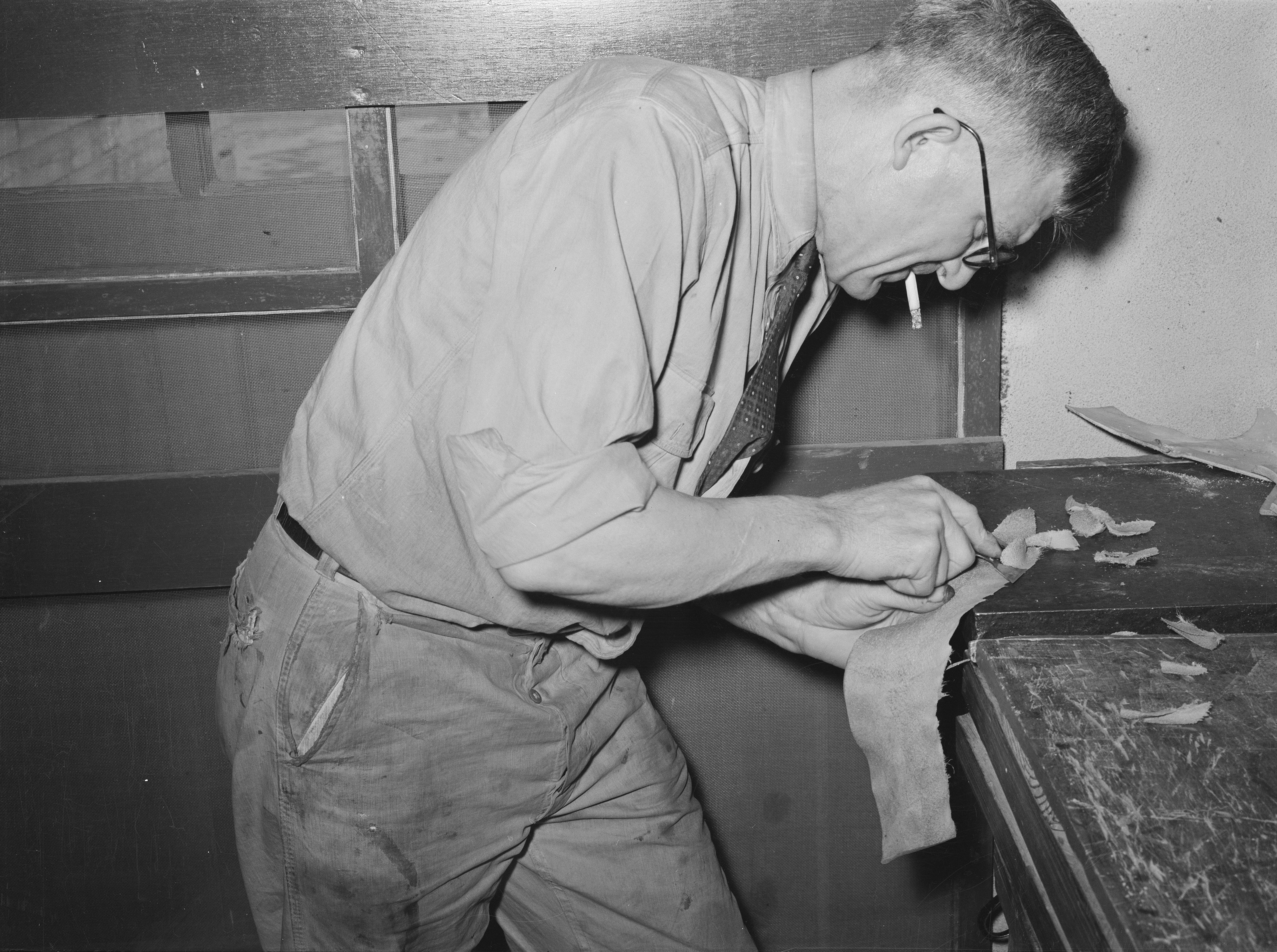A craftsman in wire-rimmed glasses bends over a wooden workbench in a Texas saddle shop, hand-skiving a strip of leather with a blade, with leather scraps and shavings visible on the bench surface.