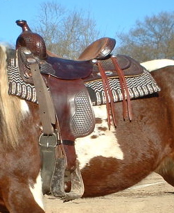 Close detail of a hand-tooled Western saddle resting on a wood rail, showing the carved leather skirt, latigo, cinch rings, and fender — components that each carry a specific name in the heritage gear vocabulary.