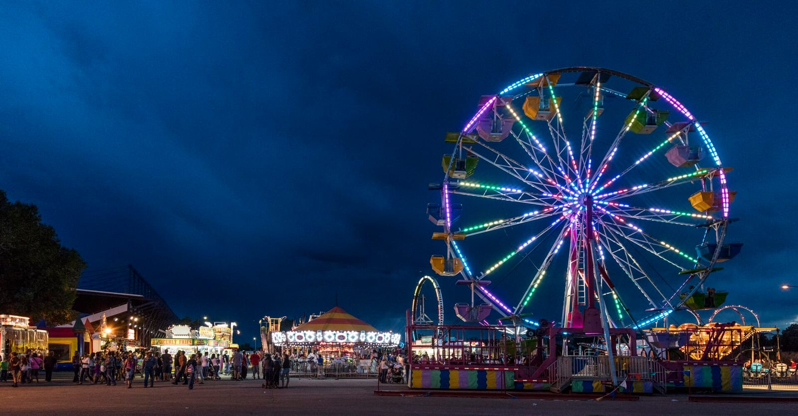 The Wyoming State Fair midway at dusk in Douglas, a lit Ferris wheel against a deep blue sky with the fairground buildings and crowd silhouetted along the row.