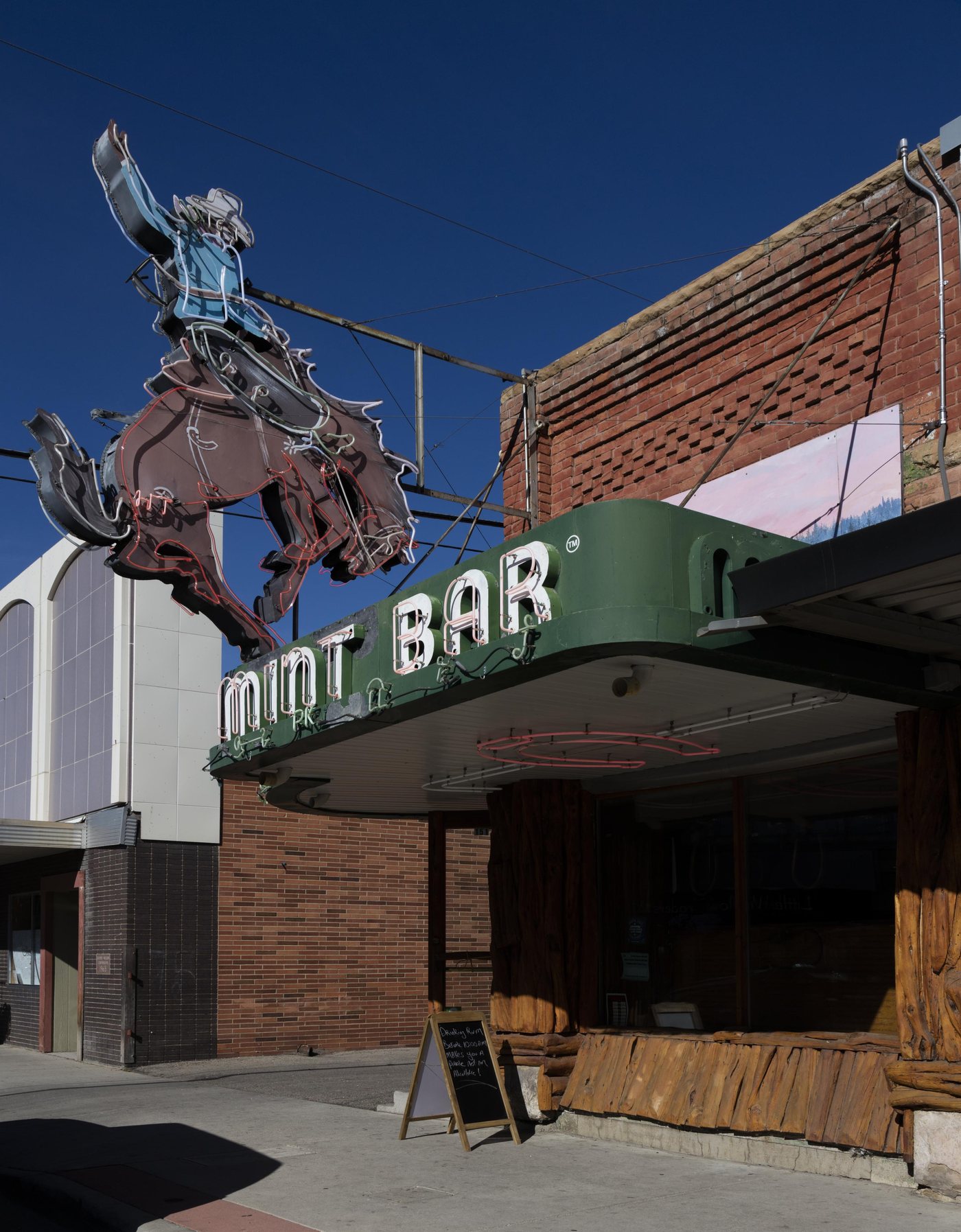 The Mint Bar on Main Street in Sheridan, its neon bucking-horse sign cantilevered over the sidewalk above a green marquee reading MINT BAR.